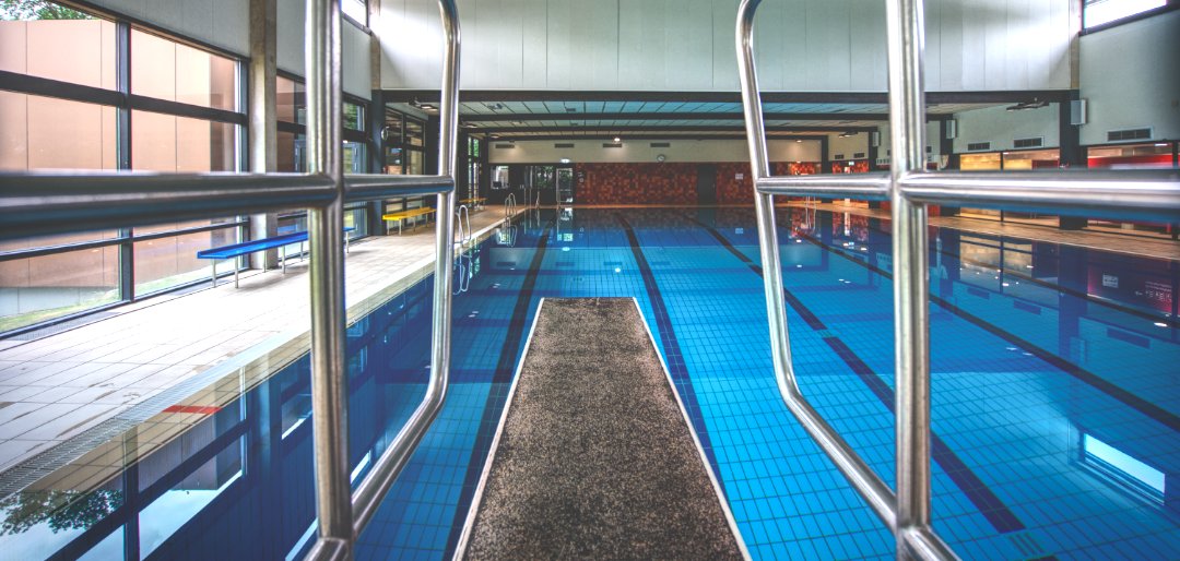 A diving board in the foreground points to the pool of an indoor swimming pool in the background