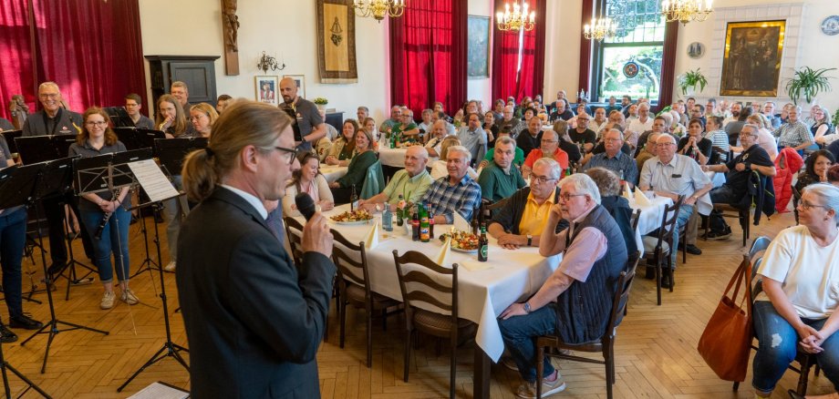 A man speaks to people sitting at tables in a hall.