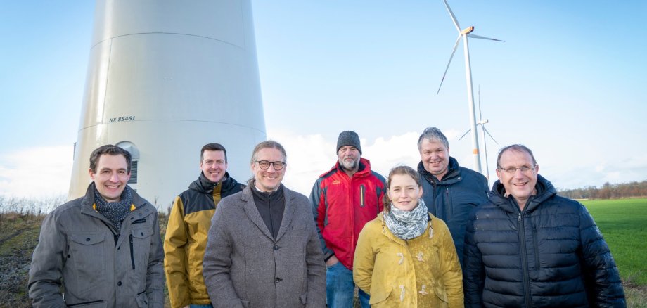 Comité van het fonds "Energie voor Kevelaer Zeven vertegenwoordigers van de fondsinvesteerders staan voor een windturbine in Kevelaer. Op de achtergrond zijn andere windturbines en een open landschap te zien. De foto symboliseert de promotie van liefdadigheidsprojecten met de opbrengst van lokale windenergie.