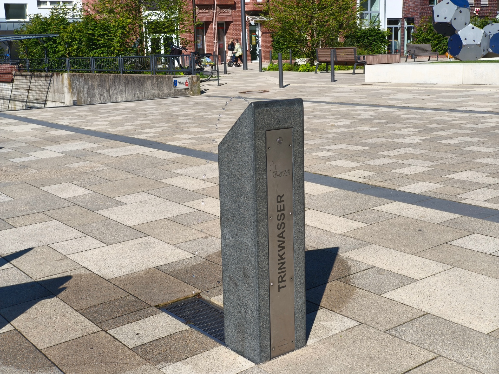 Öffentlicher Trinkwasserbrunnen aus Granit und Edelstahl auf dem Peter-Plümpe-Platz in Kevelaer. Aus dem Brunnen fließt Trinkwasser. Im Hintergrund sind Sitzbänke, Bäume und Teile des Platzes zu sehen.