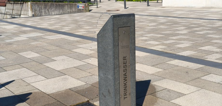 Trinkwasserbrunnen am Peter-Plümpe-Platz Öffentlicher Trinkwasserbrunnen aus Granit und Edelstahl auf dem Peter-Plümpe-Platz in Kevelaer. Aus dem Brunnen fließt Trinkwasser. Im Hintergrund sind Sitzbänke, Bäume und Teile des Platzes zu sehen.