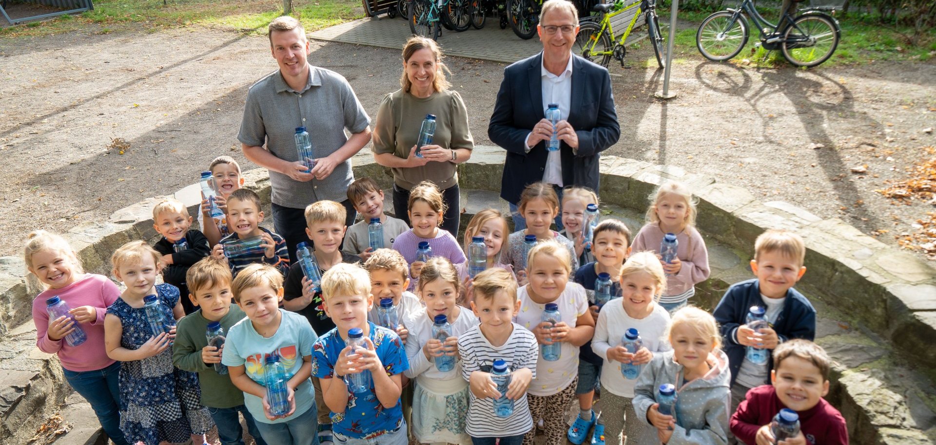 Erstklässler der St. Franziskus Grundschule in Twisteden stehen mit ihren neuen blauen Trinkflaschen auf dem Schulhof. Hinter ihnen stehen Hendrik Kruß, Rektorin Tanja Kocken und Wolfgang Toonen.