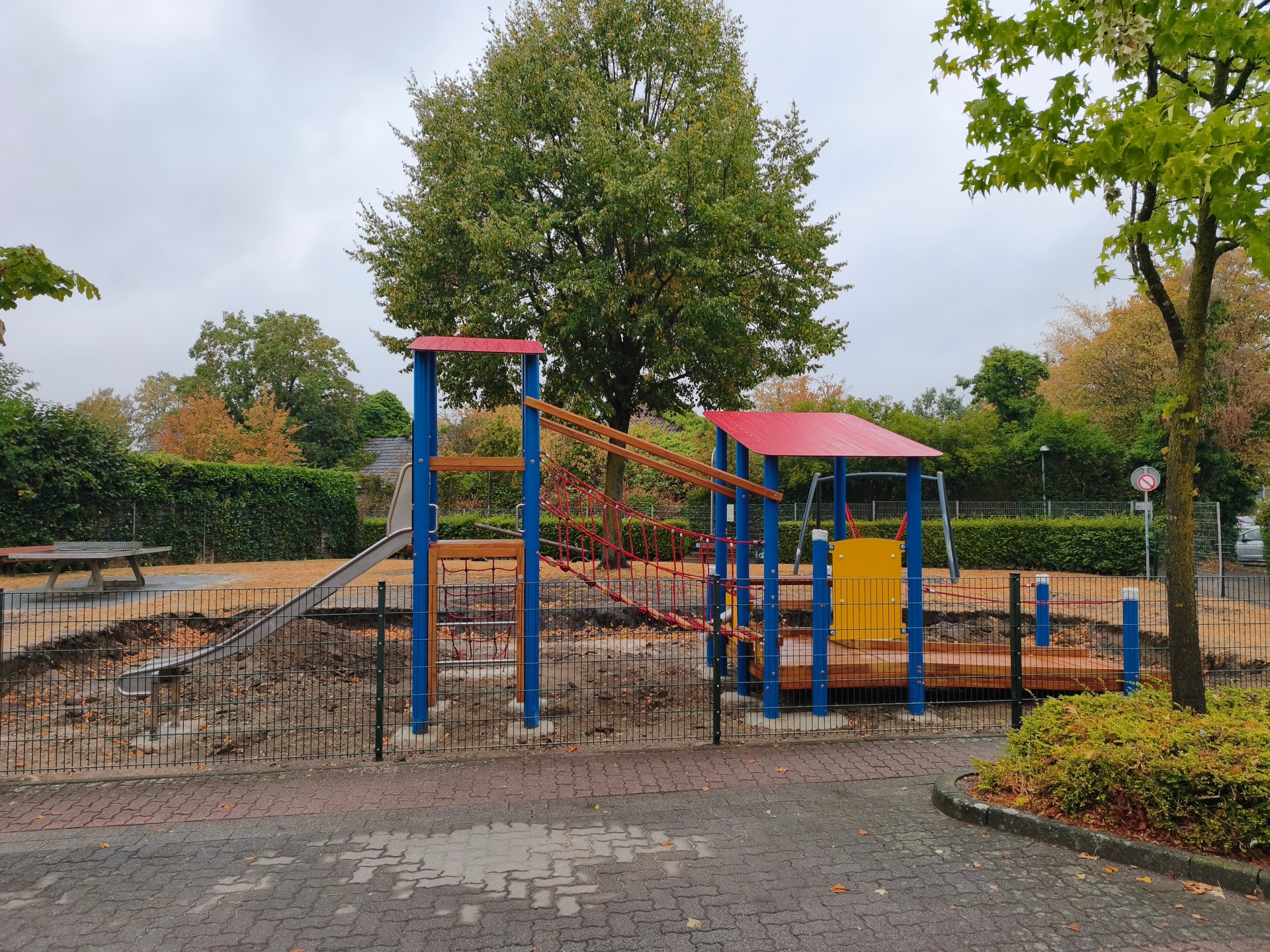 Neues blau-rot-gelbes Spielgerät auf dem Spielplatz am Theodor-Heuss-Ring mit Rutsche, Kletterelementen, Seilhängebrücke und rollstuhlgeeignetem Laufsteg.