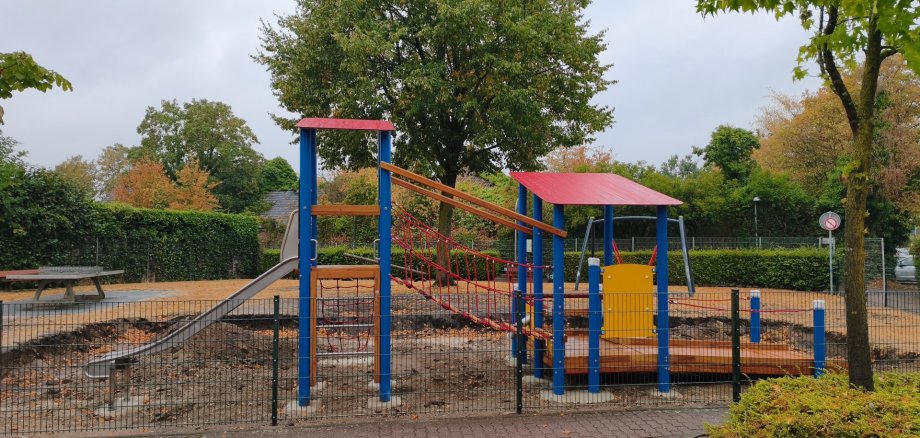Neues blau-rot-gelbes Spielgerät auf dem Spielplatz am Theodor-Heuss-Ring mit Rutsche, Kletterelementen, Seilhängebrücke und rollstuhlgeeignetem Laufsteg.