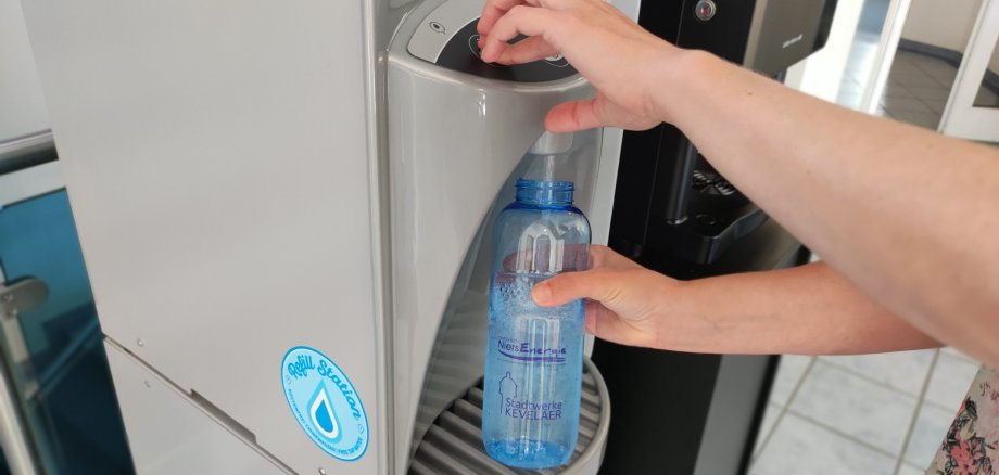 An einem Wasserspender wird eine blaue Trinkflasche gefüllt.