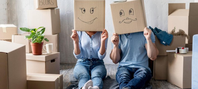 Stromvertrag beim Umzug rechtzeitig an- und abmelden Zwei Personen sitzen zwischen Umzugskartons auf dem Boden und halten sich Kartons mit aufgemalten Gesichtern vor das Gesicht. Das Bild symbolisiert einen bevorstehenden Umzug und die rechtzeitige An- oder Abmeldung des Stromvertrags.